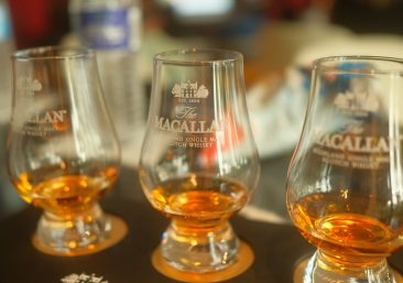 Rare Macallan Whisky Cask Ownership Single oak cask in maturation warehouse Stenciled whisky cask with distillery markings Aged Scotch whisky cask ready for investmentshowing green upward arrow; Club-Exclusive featuring premium tasting glass—all against elegant dark oak whisky warehouse texture with golden lighting.